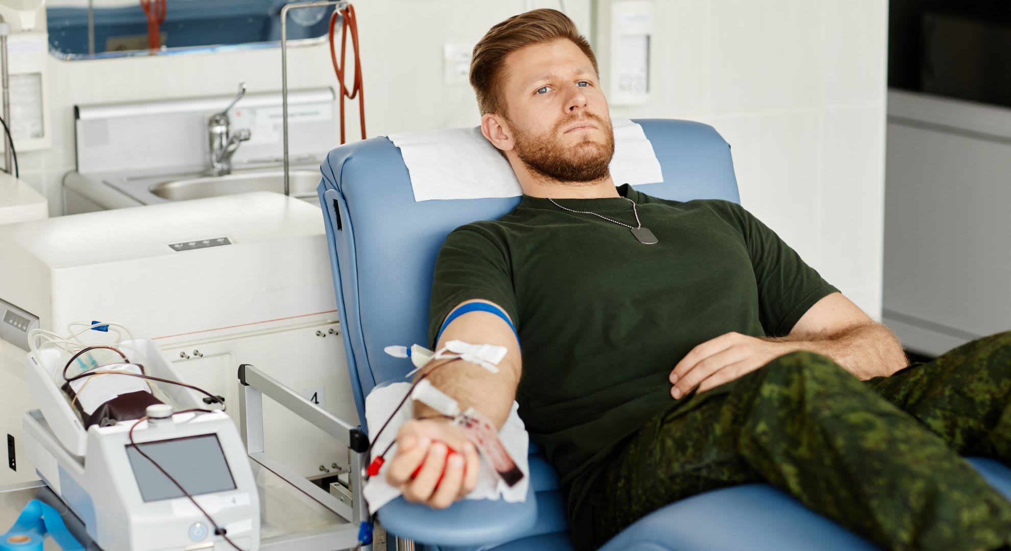 A man in military-style clothing donates blood while lying on a medical chair, similar to procedures like 10 pass ozone therapy vs EBOO therapy, with blood collection equipment beside him in a clinical setting.